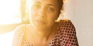 Woman sitting in soft sunlight with a calm but distant expression, appearing thoughtful and introspective.