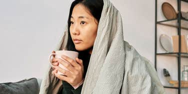 Sensitive woman sitting on a couch wrapped in a blanket, holding a mug and looking ahead in a quiet indoor setting.