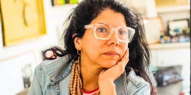 Woman with glasses resting her face in her hand, looking thoughtful while sitting indoors.