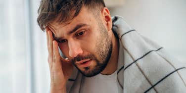 Tired man with a blanket over his shoulders holding his head and looking worn down, appearing exhausted and unwell after a long night.
