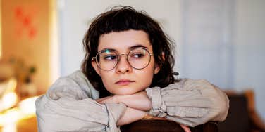 Person wearing round glasses resting their arms on a chair and looking off to the side, appearing distant and emotionally withdrawn in a softly lit room.
