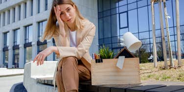 Frustrated woman sitting on bench with box of personal belongings near business center. 