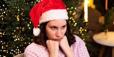 Woman in pink pajamas wearing a Santa hat, sitting in front of a Christmas tree with a defiant, fed-up expression, throwing tantrum.
