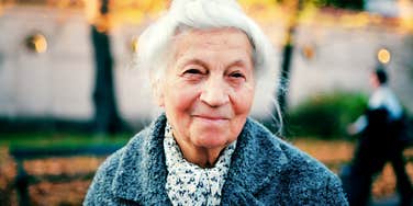 Elderly woman standing outdoors in natural light, wearing a coat and appearing calm and reflective.