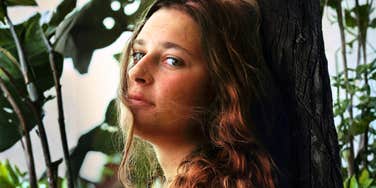 Woman standing outdoors beside a tree in natural light, looking slightly off to the side.