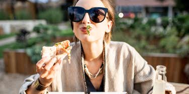 Woman wearing sunglasses enjoying food outdoors, appearing carefree, playful, and fully present in the moment.