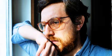 Man wearing glasses looks out a window with a thoughtful, concerned expression.