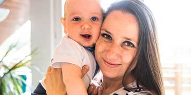 Woman holding a baby indoors near a window.