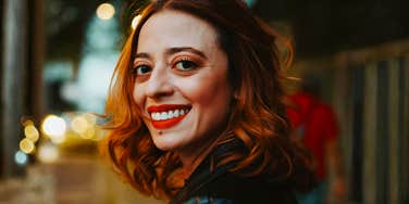 A woman with red hair smiles while turning toward the camera on a city sidewalk at dusk.