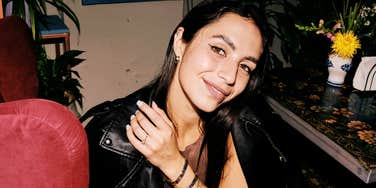 A woman sitting indoors smiling slightly while wearing a leather jacket.