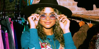 A woman wearing a black hat and sunglasses standing among clothing racks.