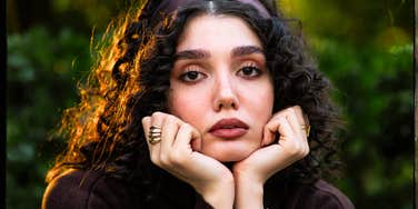A woman with curly hair rests her chin in her hands while looking directly at the camera outdoors.