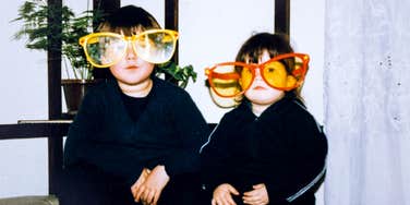 Two young children sitting indoors wearing oversized novelty sunglasses.