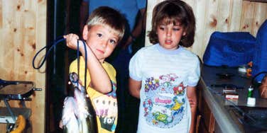 Two children standing indoors, one holding a fish on a string.