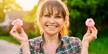 Smiling woman holding two pink flowers outdoors in natural light.