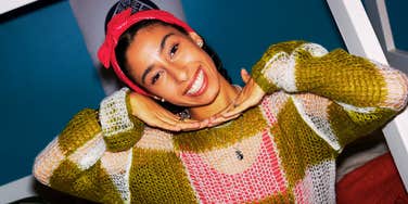 A smiling young person posing indoors with their hands under their chin.