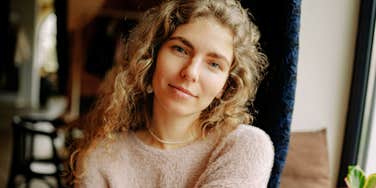 A woman with curly hair sits indoors near a window, looking calmly at the camera.