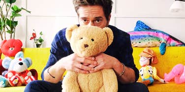 A man sitting on a couch holding a teddy bear, surrounded by stuffed toys.