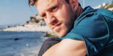 Man sitting alone near the water with a distant, contemplative expression