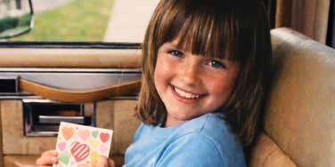 A smiling child sitting in a car seat holding a colorful card.