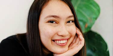 A smiling person resting their face on their hand indoors.