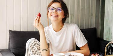 Woman wearing glasses smiling while holding a strawberry at a table with breakfast food.