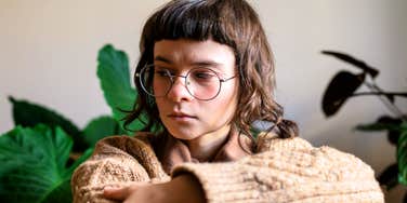 A person wearing glasses sits indoors with their arms folded, looking down thoughtfully.