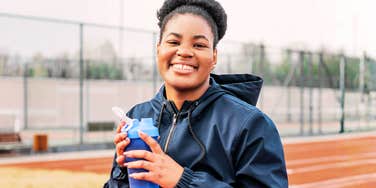 A woman smiling outdoors while holding a water bottle on a running track.
