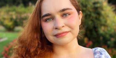 A woman with red hair looking at the camera outdoors with greenery in the background.