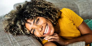 A woman smiling while resting on a couch in a relaxed home setting.