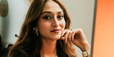 A woman sits indoors with her hand resting on her chin, looking toward the camera.