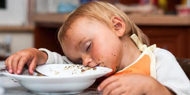 Toddler asleep at the table with food on a plate.