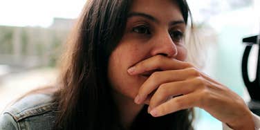A woman sitting indoors with her hand covering her mouth while looking off to the side.
