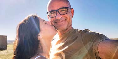 A woman kissing a man on the cheek while he takes a selfie outdoors.