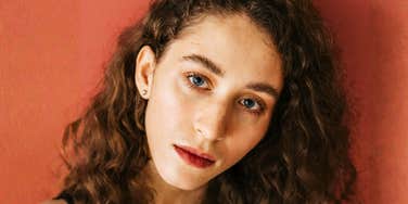 A woman with curly hair looks directly at the camera against a warm-toned background.