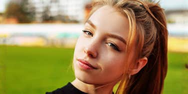 A young woman looks toward the camera while standing outdoors on a grassy field.