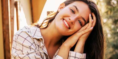 A woman smiling and resting her head on her hands outdoors in warm sunlight.