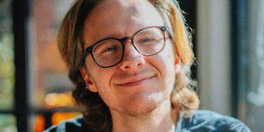A person wearing glasses smiles calmly while sitting near a window in soft natural light.