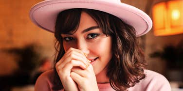 Woman wearing a hat smiles softly while looking toward the camera.