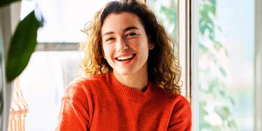 smiling woman in cozy red sweater by bright window, natural morning light on her face