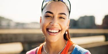A woman smiling outdoors in sunlight with buildings in the background.