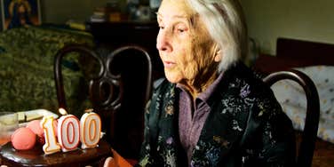 An elderly woman sitting at a table with a birthday cake topped with candles shaped like the number 100.