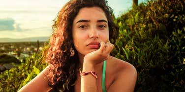 pensive young woman resting her chin on her hand at golden hour, gazing forward with distant, reflective expression against lush green backdrop