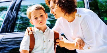 concerned mother gently holding her young son by the shoulders near a parked car during school drop-off, boy wearing a backpack with withdrawn expression, bright morning light highlighting emotional tension and parental worry