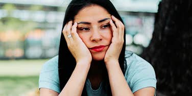 Young woman sitting on a park bench, resting her face in her hands with a pensive, melancholic expression, appearing lost in thought.