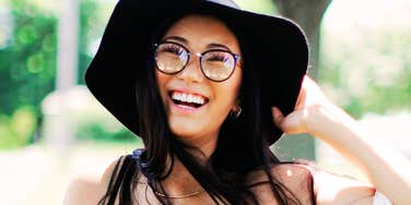 Smiling woman wearing glasses and a wide-brim hat outdoors, laughing in natural sunlight.