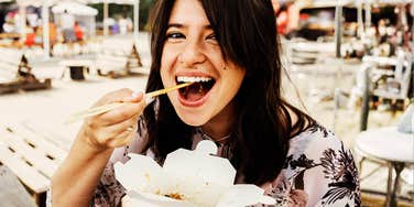 close-up of smiling young woman eating noodles with chopsticks from a takeout container at an outdoor market