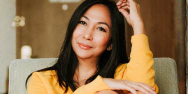Woman in a yellow sweater sitting on a chair indoors, resting her head on her hand with a calm, reflective expression.