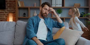 dad stressed out sitting on couch while daughter runs behind him