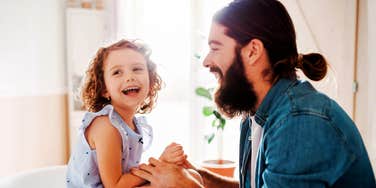 A father smiling and playing with his young child in a bright room.